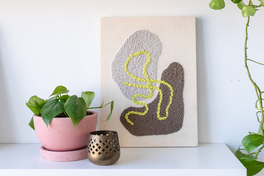 The Pebbles II punch needle design features abstract taupe and linen shapes with a neon citron 'squiggle', and is posed on a white shelf with a small houseplant.