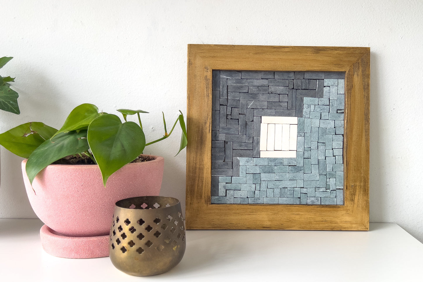 Framed mosaic mirror on a white surface with a plant and candle holder.