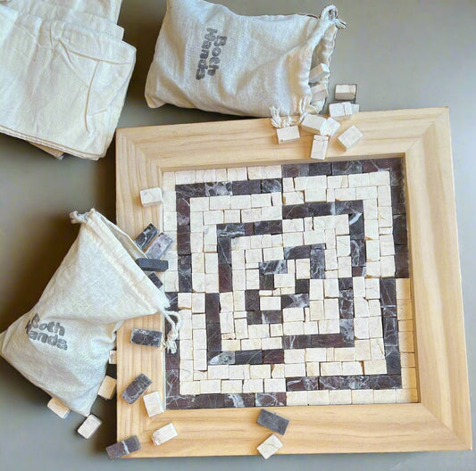 A mosaic kit in progress with tiles laid out to form a geometric pattern on a wooden frame, alongside two bags of tiles and additional tiles scattered around.
