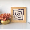 The Offset mosaic design, which has a wood frame, beige and rosso levanto marble tiles in an optical illusion design, sits on a white shelf with fresh flowers.