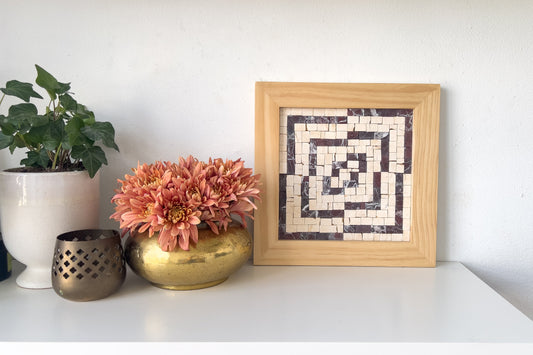 The Offset mosaic design, which has a wood frame, beige and rosso levanto marble tiles in an optical illusion design, sits on a white shelf with fresh flowers.
